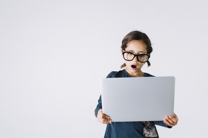 Dziewczynka w za dużych okularach na tle szarej ściany trzyma laptop z zdziwionym wyrazem twarzy