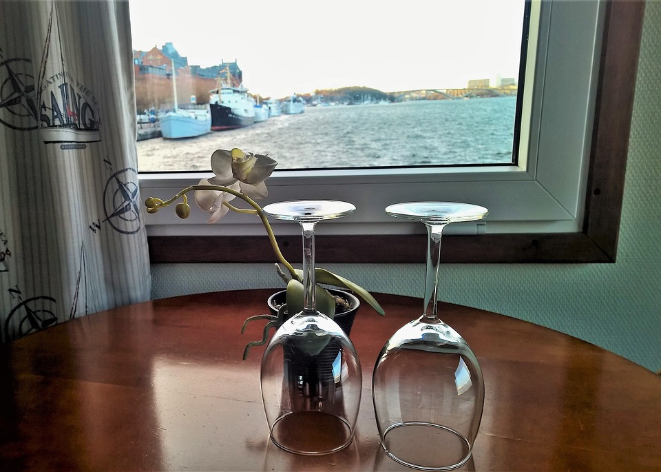 Dwie lampki do wina odwrócone do góry stopką stojące na polakierowanym drewnianym stole, obok kwiatka w doniczce na tle okna z widokiem na szeroką rzekę, statki i most
