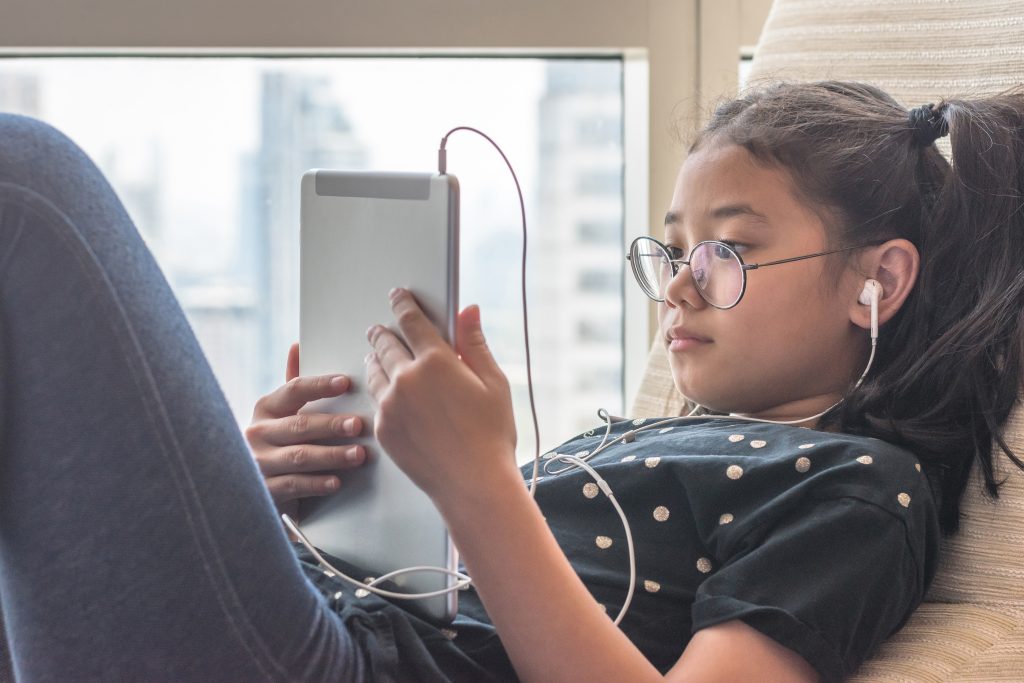 Dziewczynka leżąca przy oknie oglądająca film na tablecie z podłączonymi do niego słuchawkami dousznymi
