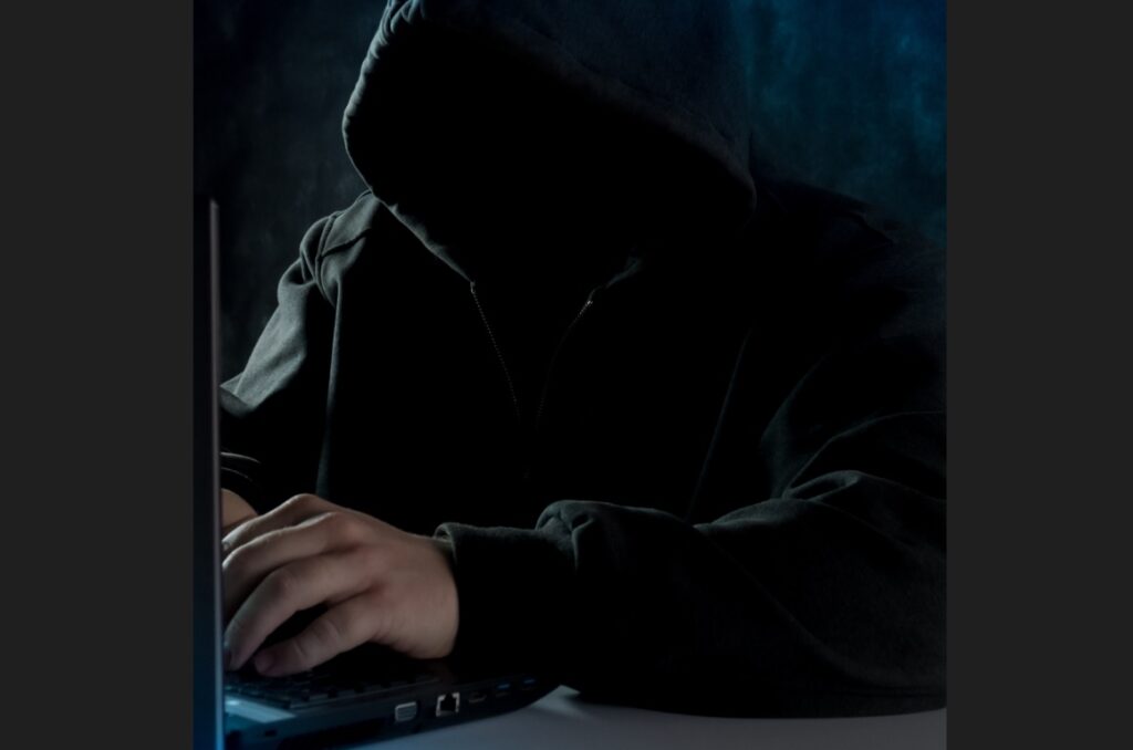 człowiek z zasłoniętą kapturem twarzą przy otwartym laptopie w ciemnym pomieszczeniu