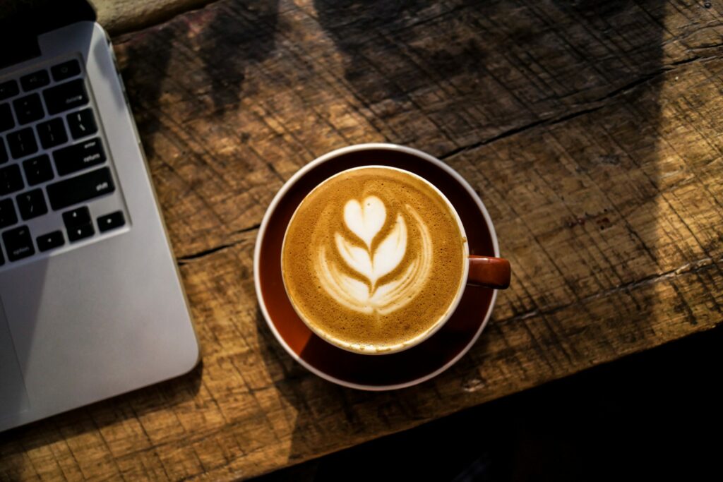 na tle ładnego brązowego blatu kawałek otwartego laptopa i filiżanka cappuccino, na powierzchni którego jest kwiatek
